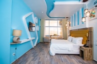 A modern hotel room featuring a minimalist design with a blue and white palette. The room has a comfortable-looking bed with white linens and a yellow headboard. A window at the far end of the room lets in natural light and offers a view of the city. Decorative ceiling lights hang above, and the walls have geometric shapes for added design flair. A wall-mounted flat-screen TV and a contemporary lamp sit on minimalist shelves.