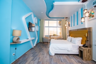 A modern hotel room featuring a minimalist design with a blue and white palette. The room has a comfortable-looking bed with white linens and a yellow headboard. A window at the far end of the room lets in natural light and offers a view of the city. Decorative ceiling lights hang above, and the walls have geometric shapes for added design flair. A wall-mounted flat-screen TV and a contemporary lamp sit on minimalist shelves.
