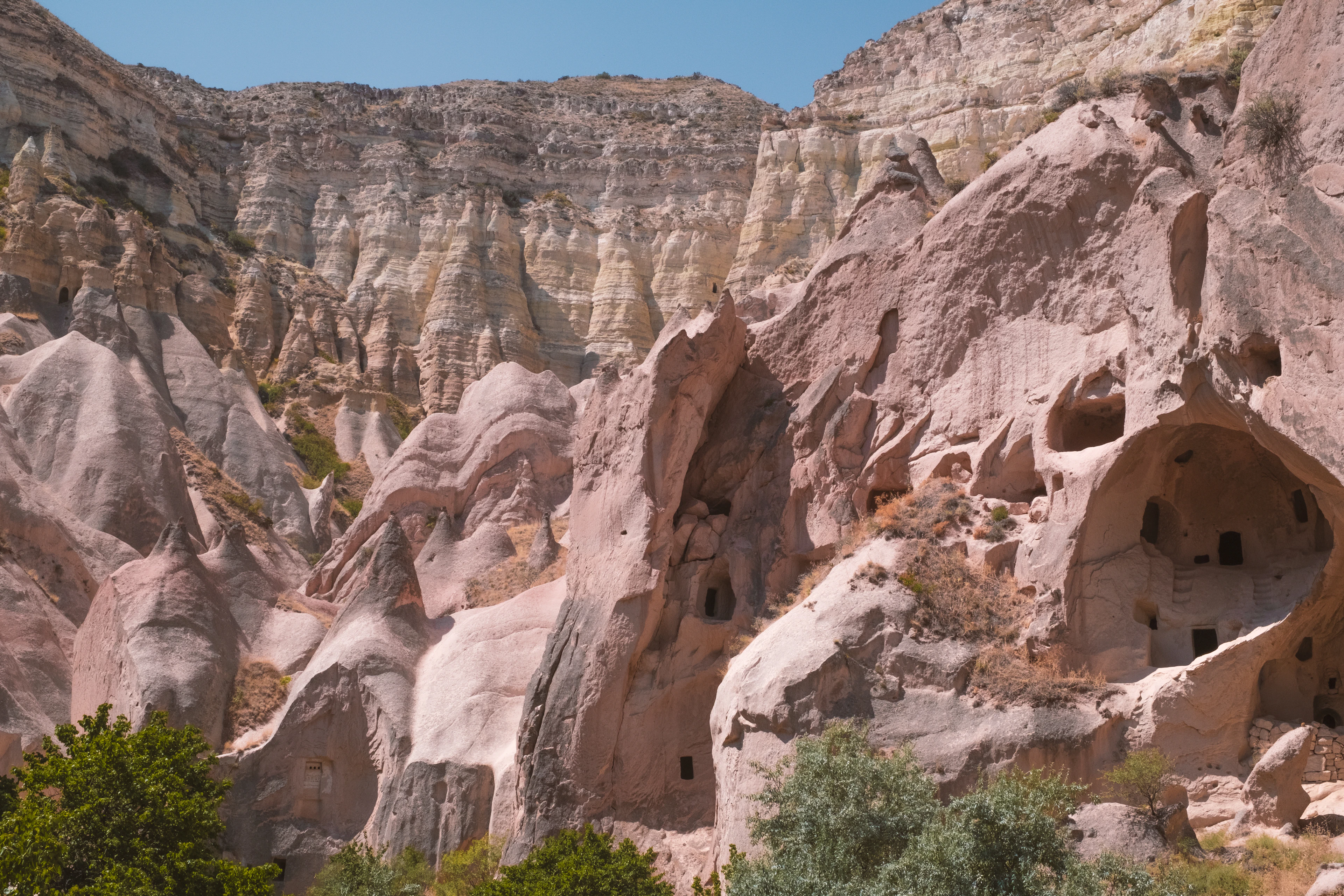 Pink-toned Rose Valley cliffs riddled with carved cave openings under a clear blue sky.