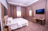 A neatly arranged hotel room with a large bed covered in white linens. The room features a light brown and beige color scheme with matching curtains. There is a flat-screen television mounted on the wall, a wooden desk with amenities such as a kettle and cup, and a window letting in natural light.