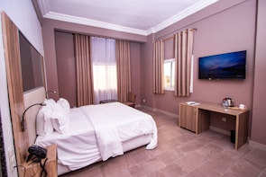 A neatly arranged hotel room with a large bed covered in white linens. The room features a light brown and beige color scheme with matching curtains. There is a flat-screen television mounted on the wall, a wooden desk with amenities such as a kettle and cup, and a window letting in natural light.