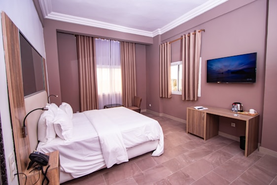A neatly arranged hotel room with a large bed covered in white linens. The room features a light brown and beige color scheme with matching curtains. There is a flat-screen television mounted on the wall, a wooden desk with amenities such as a kettle and cup, and a window letting in natural light.