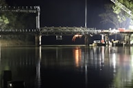 A partially constructed bridge spans over a calm body of water at night. Bright lights illuminate the construction site, casting reflections in the water. Sparks are visible from welding or construction activity on the bridge.