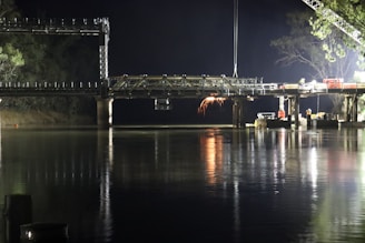 A partially constructed bridge spans over a calm body of water at night. Bright lights illuminate the construction site, casting reflections in the water. Sparks are visible from welding or construction activity on the bridge.