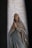 A detailed sculpture of a religious figure in a praying posture with hands clasped together. The figure is draped in a robe with blue and white colors, standing against a background featuring inscribed names and a cross symbol.