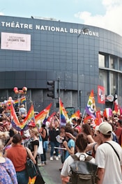 A lively outdoor scene with a crowd of people participating in a demonstration or parade, holding and waving colorful flags with rainbow and red designs. The event is taking place in front of a modern building labeled 'THÉÂTRE NATIONAL BRETAGNE'. Attendees seem engaged and there is a sense of celebration and advocacy.