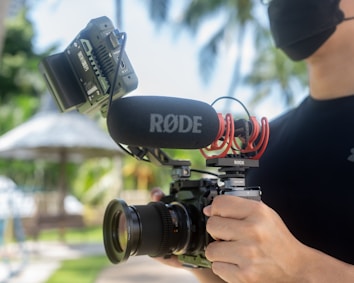A person holds a professional video camera equipped with a large lens and a R&Oslash;DE microphone. The person is wearing a black face mask and a dark shirt. The camera has a mounted external monitor. The background features blurred greenery and palm trees, suggesting an outdoor setting.
