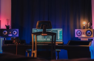 A modern music studio setup featuring a chair positioned in front of a desk with a computer monitor displaying audio editing software. Large speakers are placed on either side of the desk, and there is ambient lighting in shades of blue and pink. A pair of headphones rests on the left side of the desk, and a dimly lit bulb is visible on the right.