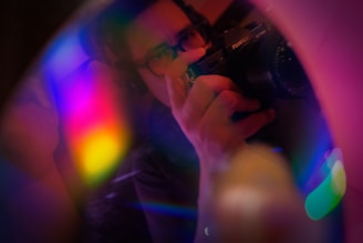 A person is holding a camera, with a lens focused on their reflection. Vibrant, multicolored light reflections fill the foreground, creating a dreamy and artistic effect.