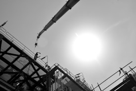 A black and white image featuring construction scaffolding and a crane arm extending diagonally across the top. A person is visible wearing safety gear, working on the structure against a bright, glaring sun in the sky.