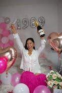 A person is celebrating joyfully with a champagne bottle and a glass in hand. The room is decorated with numerous pink, white, and metallic balloons. In the background, there are large metallic balloons forming the number '1989'. The person is sitting on a bed which is covered with a pink and white bedspread, and there are heart-shaped balloons and a bouquet of flowers visible.