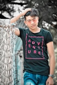 A young man wearing a black t-shirt with the word 'ADVENTURE' in pink letters, blue jeans, and pink-tinted sunglasses is posing casually with one hand in his hair. He is standing next to a metal fence with a blurry outdoor background of trees.