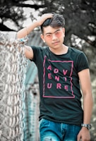 A young man wearing a black t-shirt with the word 'ADVENTURE' in pink letters, blue jeans, and pink-tinted sunglasses is posing casually with one hand in his hair. He is standing next to a metal fence with a blurry outdoor background of trees.