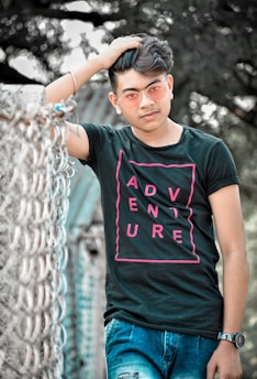A young man wearing a black t-shirt with the word 'ADVENTURE' in pink letters, blue jeans, and pink-tinted sunglasses is posing casually with one hand in his hair. He is standing next to a metal fence with a blurry outdoor background of trees.