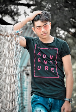 A young man wearing a black t-shirt with the word 'ADVENTURE' in pink letters, blue jeans, and pink-tinted sunglasses is posing casually with one hand in his hair. He is standing next to a metal fence with a blurry outdoor background of trees.