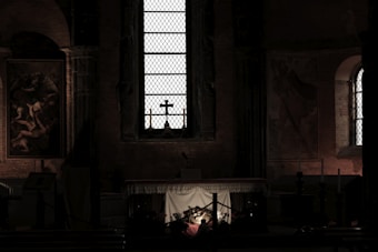 A dimly lit church interior features a prominent stained glass window at the center. Below the window is an altar with a cross silhouetted against it. The surrounding walls are adorned with religious paintings and dim, ambient light highlights some architectural details.