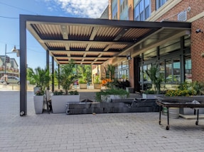 A modern outdoor patio area with a metal pergola, surrounded by large planters filled with lush green palm plants and bright flowers. The setting is adjacent to a brick and glass building, with a wide paved walkway in the foreground. The sky is partly cloudy.