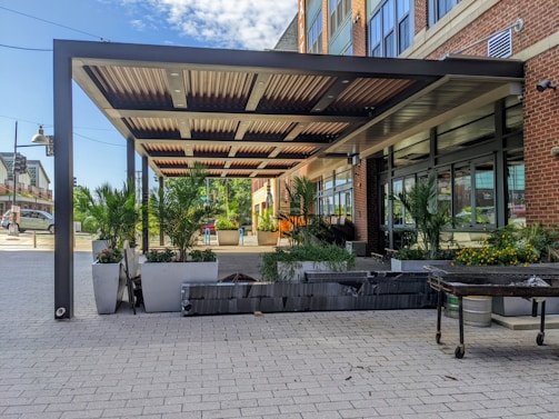 A modern outdoor patio area with a metal pergola, surrounded by large planters filled with lush green palm plants and bright flowers. The setting is adjacent to a brick and glass building, with a wide paved walkway in the foreground. The sky is partly cloudy.