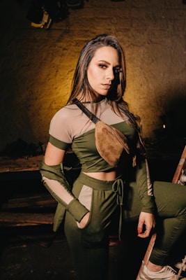 A woman with long hair and makeup wearing a stylish green and beige outfit and a brown crossbody bag. She is posing with one leg on a step or platform in a dimly lit environment with a textured wall in the background.