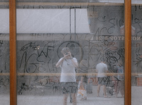 A person is reflected in a large glass pane, covered in layers of writing and scribbles. The surface features various abstract shapes and words, with some areas slightly more opaque due to the text. Behind the glass, two people are partially visible, adding depth to the scene.