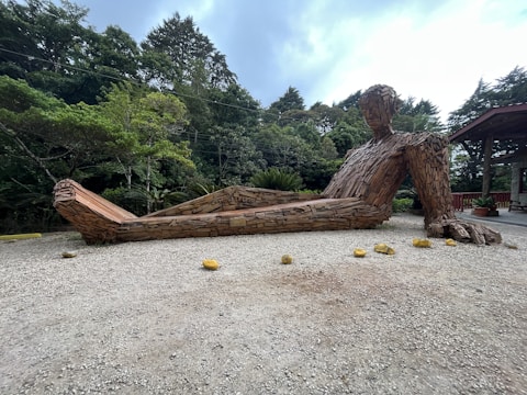 A large wooden sculpture of a reclining human figure is situated outdoors on a gravel surface, surrounded by lush green trees. The sculpture is made from stacked pieces of wood, creating a textured appearance. The environment appears natural and serene, with a wooden structure visible on the right side.