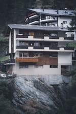 A multi-story house built on a rocky hillside, featuring wooden and concrete exterior elements. The structure includes balconies and large windows, integrating natural wood tones with modern architectural features. The surrounding area includes trees and vegetation, adding to the secluded mountain environment.