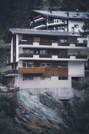 A multi-story house built on a rocky hillside, featuring wooden and concrete exterior elements. The structure includes balconies and large windows, integrating natural wood tones with modern architectural features. The surrounding area includes trees and vegetation, adding to the secluded mountain environment.
