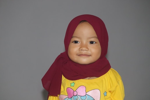 A young child wearing a maroon hijab and a yellow shirt with a colorful design on the front. The background is plain gray, and the child is smiling gently.