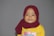 A young child wearing a maroon hijab and a yellow shirt with a colorful design on the front. The background is plain gray, and the child is smiling gently.