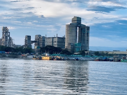 A large industrial factory complex sits on the riverside, featuring tall silos and various industrial buildings. The reflection of the structures is visible in the calm water, with several boats docked near the shore. The sky is partly cloudy, adding a tranquil and serene backdrop to the scene.