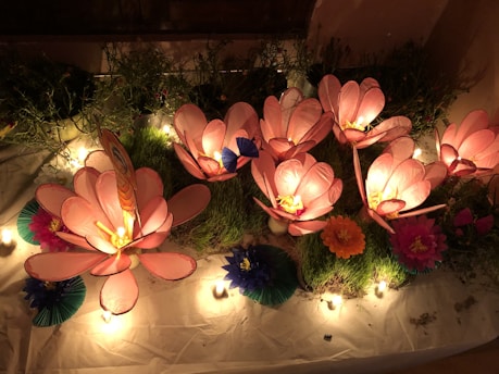 Large, illuminated pink flowers are arranged on a bed of green grass. The flowers are surrounded by small, colorful paper decorations and warm, glowing lights, creating a whimsical garden scene.