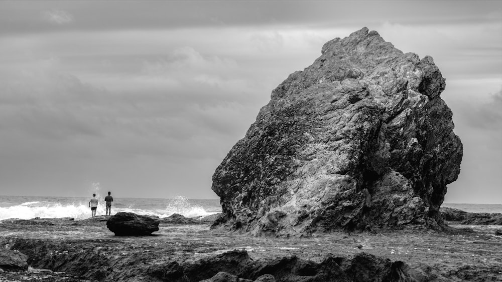 a couple of people standing next to a large rock on a beach