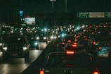A busy highway at night with a dense flow of vehicles, their headlights and taillights illuminating the scene. Overhead signage can be seen in the background, with various traffic signs and advertisements partially visible. The overall scene conveys a sense of congestion and urban nightlife.