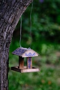 A wooden birdhouse hangs from a tree branch, adorned with blue floral designs and the word 'Lowe's' on it. The backdrop is a blurred, lush green garden.