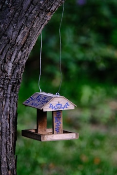 A wooden birdhouse hangs from a tree branch, adorned with blue floral designs and the word 'Lowe's' on it. The backdrop is a blurred, lush green garden.