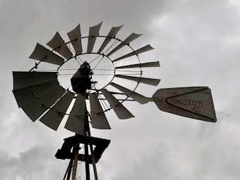 A large, metal windmill with a distinctive circular rotor featuring multiple blades stands against a cloudy sky. The structure is aged, and the tail vane displays the words 'The Aermotor Chicago.' The backdrop is a muted gray, suggesting overcast weather.
