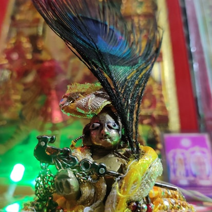 A detailed close-up of a small, intricately decorated figurine wearing vibrant golden and colorful garments. The figure has an elaborate peacock feather headdress. The background features blurred bright colors and festive lights, suggesting a decorative or celebratory setting.