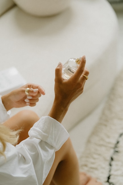 A person with a light skin tone holding a clear perfume bottle labeled 'Gisou' in a relaxed indoor setting. The person is wearing a white shirt and has neatly manicured nails. The background includes a plush cream-colored rug and minimalistic furniture.