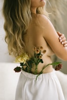 A woman with long, wavy blonde hair is partially nude from the back, with her arm crossed over her chest. In her loose white pants, flowers of different colors are tucked in, creating a contrast with her skin tone. The setting seems serene and natural, emphasizing a connection with nature.