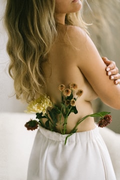 A woman with long, wavy blonde hair is partially nude from the back, with her arm crossed over her chest. In her loose white pants, flowers of different colors are tucked in, creating a contrast with her skin tone. The setting seems serene and natural, emphasizing a connection with nature.