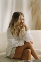 A person with long blonde hair sits on a light-colored sofa, wearing a loose white shirt. The setting appears minimalist, with soft, neutral tones. The person's expression is contemplative, with one hand resting on their face.