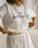 A person wearing a white t-shirt with 'Earth Day' and a small flower design printed on it is slightly lifting the shirt. The shirt has a casual style and the lower garment is also white, creating a monochrome look. The setting appears soft and minimalistic, likely indoors.