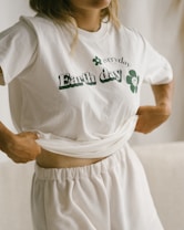 A person wearing a white t-shirt with 'Earth Day' and a small flower design printed on it is slightly lifting the shirt. The shirt has a casual style and the lower garment is also white, creating a monochrome look. The setting appears soft and minimalistic, likely indoors.