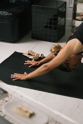 A person is practicing yoga in a downward-facing position on a black mat. Nearby, there are small stacked objects that appear to be yoga blocks. The setting includes a black leather sofa and a modern black tiled cube, suggesting an indoor space with a calm atmosphere.