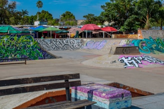 A vibrant skate park adorned with colorful graffiti art covers various structures including ramps and benches. In the background, several outdoor canopies in bright colors provide shade. The area is surrounded by lush green trees under a clear blue sky.