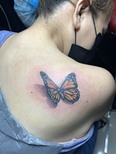 A person is sitting with their back turned, displaying a freshly inked butterfly tattoo on their shoulder. The butterfly has vibrant orange wings with black patterns and fine white dots along the edges. The skin around the tattoo appears slightly red, suggesting it is newly done. The person has light brown hair tied back, and is wearing a surgical mask and a blue hair cover, indicating they are likely in a professional tattoo setting.