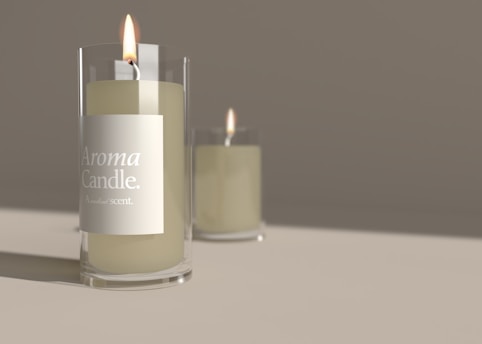 A close-up view of an elegant scented candle placed in a transparent glass holder, with the words 'Aroma Candle. A natural scent.' displayed on the label. Another similar candle is visible in the blurred background, enhancing the atmosphere.