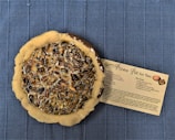 A pecan pie with a partially baked crust is placed on a blue textured surface. Next to the pie is a recipe card titled 'Pecan Pie for Two' detailing the ingredients and instructions for making the pie. The pie appears glossy with a rich filling of pecans and syrup.