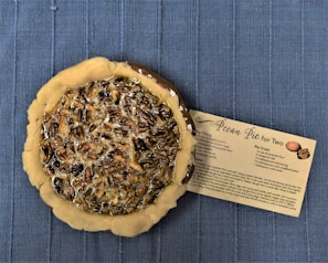 A pecan pie with a partially baked crust is placed on a blue textured surface. Next to the pie is a recipe card titled 'Pecan Pie for Two' detailing the ingredients and instructions for making the pie. The pie appears glossy with a rich filling of pecans and syrup.