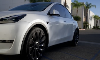 A sleek white car with modern design elements, featuring stylish black wheels. It is parked in a sunlit urban environment with palm trees visible in the background. The car's front and side are prominently displayed, highlighting its smooth curves and tinted windows.